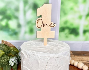May include: A round cake covered in white textured frosting, topped with a light-colored wooden cake topper shaped like the number one with the word One written in cursive script. A small wooden sign with the text decor by luciana is present.