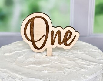 May include: A light-colored wooden cake topper featuring the word One in a dark brown cursive script font. The topper has a single vertical stake extending from the bottom, which is inserted into a round cake covered in smooth white frosting.