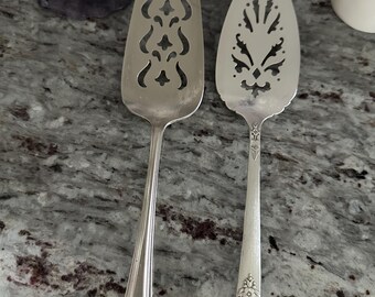 May include: Two silver cake servers with ornate cutouts. The servers have decorative handles with floral designs.