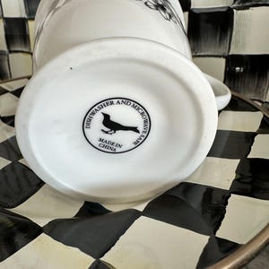 May include: White ceramic mug with a black and white floral design. The bottom of the mug has a black bird logo and text that reads "Dishwasher and Microwave Safe" and "Made in China". The mug is on a black and white checkered plate.