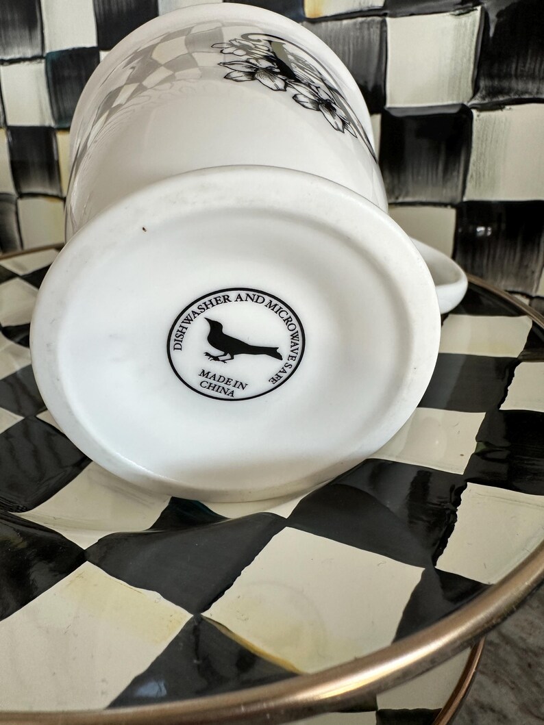 May include: White ceramic mug with a black and white floral design. The bottom of the mug has a black bird logo and text that reads "Dishwasher and Microwave Safe" and "Made in China". The mug is on a black and white checkered plate.