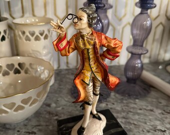 May include: A figurine of a man in a red and yellow suit holding a pair of spectacles. The figurine is standing on a black marble base.