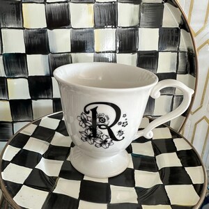 May include: A white teacup with a black floral design and the letter "R" is centered on a black and white checkered plate. The teacup has a handle and sits on a pedestal base. A larger checkered plate is behind the teacup.