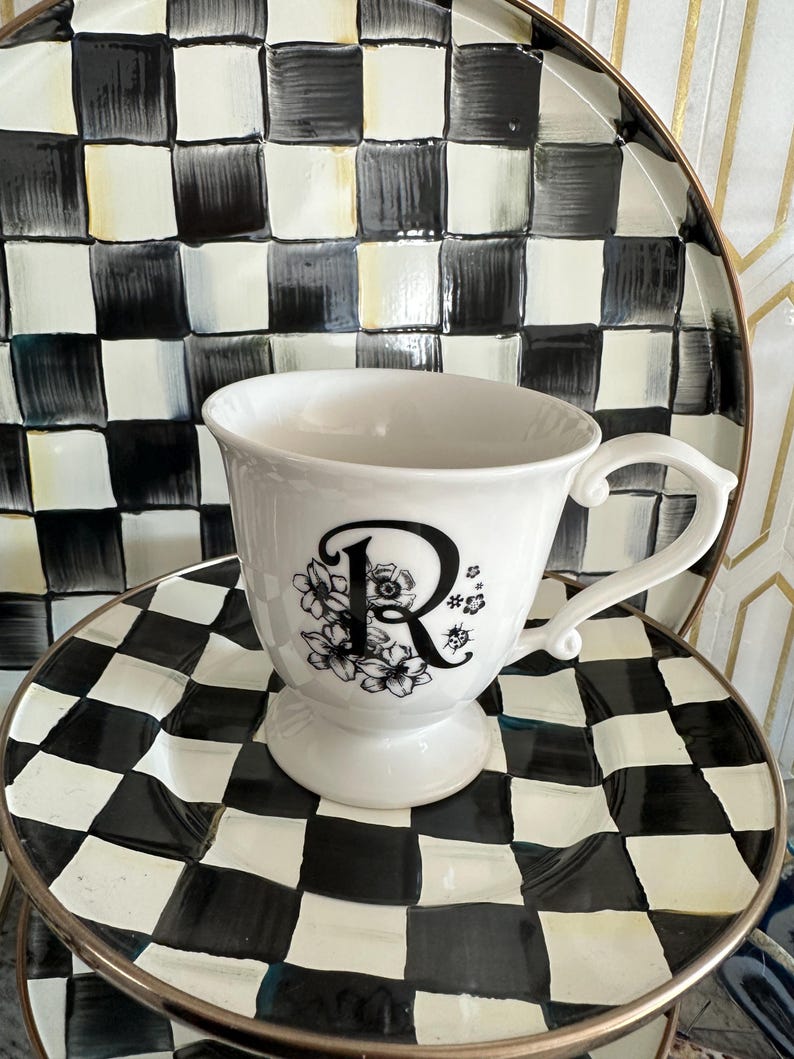 May include: A white teacup with a black floral design and the letter "R" is centered on a black and white checkered plate. The teacup has a handle and sits on a pedestal base. A larger checkered plate is behind the teacup.
