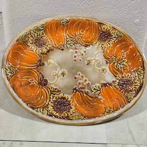Puede incluir: Una bandeja de servicio ovalada de cerámica con un borde en relieve de calabazas naranjas, girasoles marrones y pequeñas bayas rojas. El centro de la bandeja tiene un acabado esmaltado en color beige claro con detalles de enredaderas.