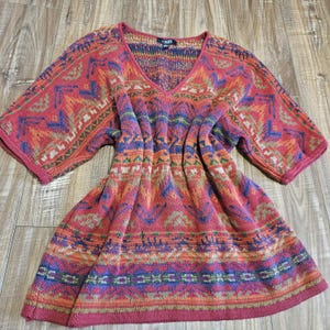 May include: A short-sleeved knit sweater featuring a V-neckline and a vibrant, multi-colored geometric pattern in shades of red, blue, orange, and green. The garment includes a Chaps brand label at the inner neckline and has a ribbed hem.