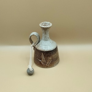 May include: A small, brown and white ceramic jug with a handle and a gray stone attached to a string. The jug has a textured surface and a carved design.