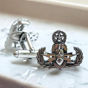 May include: A pair of silver-toned metal cufflinks featuring a detailed military-style insignia. The design includes a central bomb motif with a star, flanked by lightning bolts and laurel wreaths, topped with a small star within a circular wreath.