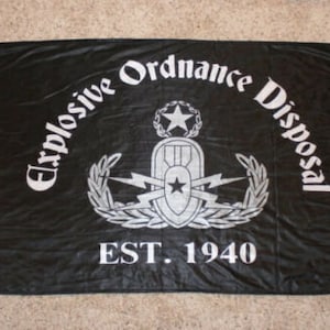 May include: A black flag with the words "Explosive Ordnance Disposal" in white lettering, a white star and a white insignia with lightning bolts, and the text "EST. 1940" in white.