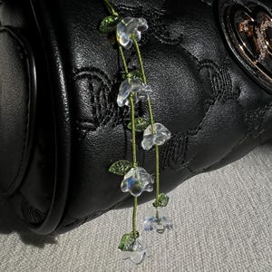 Può includere: Una borsa in pelle nera trapuntata con un ciondolo floreale decorativo. Il ciondolo presenta perline trasparenti a forma di fiore infilate su un filo verde, con piccoli accenti di foglie verdi. La borsa ha un'emblema color oro.
