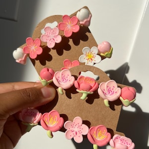 May include: A selection of pink and white floral accessories, possibly hair clips or embellishments, presented on a brown cardboard display. The flowers have varied designs, some with multiple petals and yellow centres.