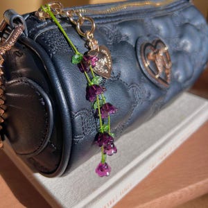 May include: Navy blue quilted handbag with a gold zip and heart-shaped emblem. A decorative charm with purple flower beads and a gold heart pendant hangs from the zip. The bag is resting on a book.