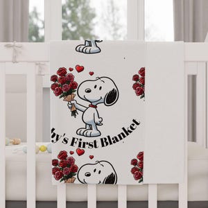 Puede incluir: Manta de bebé blanca con Snoopy sosteniendo un ramo de rosas rojas y corazones rojos. El texto "Baby's First Blanket" está impreso en la manta. La manta está doblada sobre una cuna.