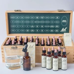 May include: A wooden box set with multiple small brown bottles with pink droppers. A glass of water and a single bottle with a dropper are in front of the box. The box has a green interior with labeled spaces for the bottles. The bottles have white labels.