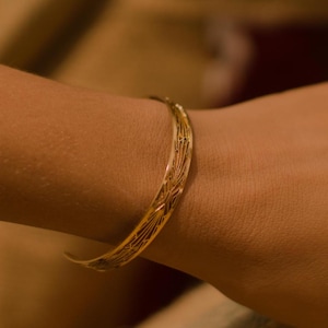 Peut inclure: Un bracelet jonc en métal doré orné d'un motif linéaire délicatement gravé sur toute sa surface. Ce bijou présente une finition polie et lisse ainsi qu'une forme incurvée ouverte qui repose sur le poignet, mettant en valeur ses détails fins.