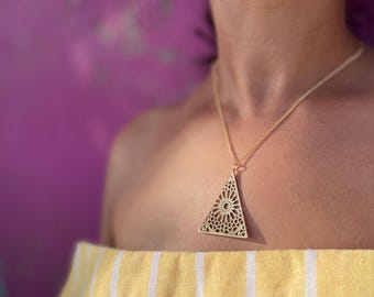 May include: A gold-toned necklace featuring a triangular pendant with an intricate, openwork geometric pattern. The pendant hangs from a delicate chain, resting against a yellow and white vertically striped garment.