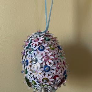 May include: An egg-shaped ornament covered in white daisy-shaped decorations with colorful sequin centers. The ornament is adorned with small, iridescent beads and has a light blue ribbon for hanging. The background is a solid, light yellow color.