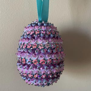 May include: An egg-shaped ornament covered in pink, purple, and silver sequins and beads. A turquoise ribbon is attached at the top for hanging. The ornament is likely for Easter or general home decor.