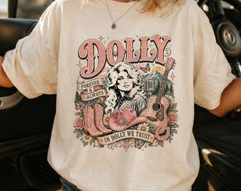May include: An off-white t-shirt featuring a graphic print of a woman with blonde hair, a guitar, cowboy boots, and a hat. The text reads DOLLY, FOREVER & ALWAYS, and IN DOLLY WE TRUST. The design includes floral accents, butterflies, and mountains.