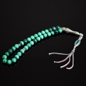 May include: A string of prayer beads featuring round, banded green malachite stones. The beads are accented with small, intricate silver-toned metal spacers and terminate in an ornate silver-toned metal tassel with four dangling chains.