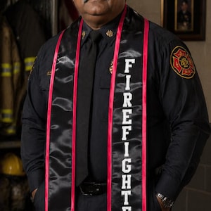 Può includere: Una persona in uniforme nera con cravatta nera e una fascia con la scritta "FIREFIGHTER" in lettere bianche con bordo rosso. La persona indossa un orologio e ha i baffi.