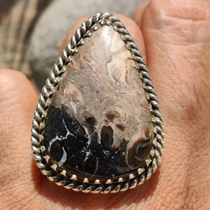 May include: A teardrop-shaped ring featuring a polished stone with a mix of brown, black, and white patterns. The stone is set in a silver-colored metal with a twisted rope design around the edge. The ring is a unique piece of jewelry.