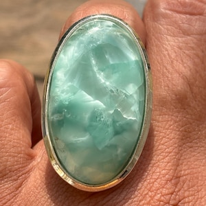 May include: An oval-shaped ring featuring a large, polished, light blue and white marbled stone set within a smooth, silver-toned metal bezel. The stone displays a translucent, crystalline texture with varying shades of pale turquoise and white.