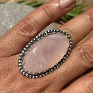 May include: Oval rose quartz ring with a silver beaded border. The large, polished pink stone is set in a detailed, textured band. The ring is a statement piece, showcasing the natural beauty of the gemstone.