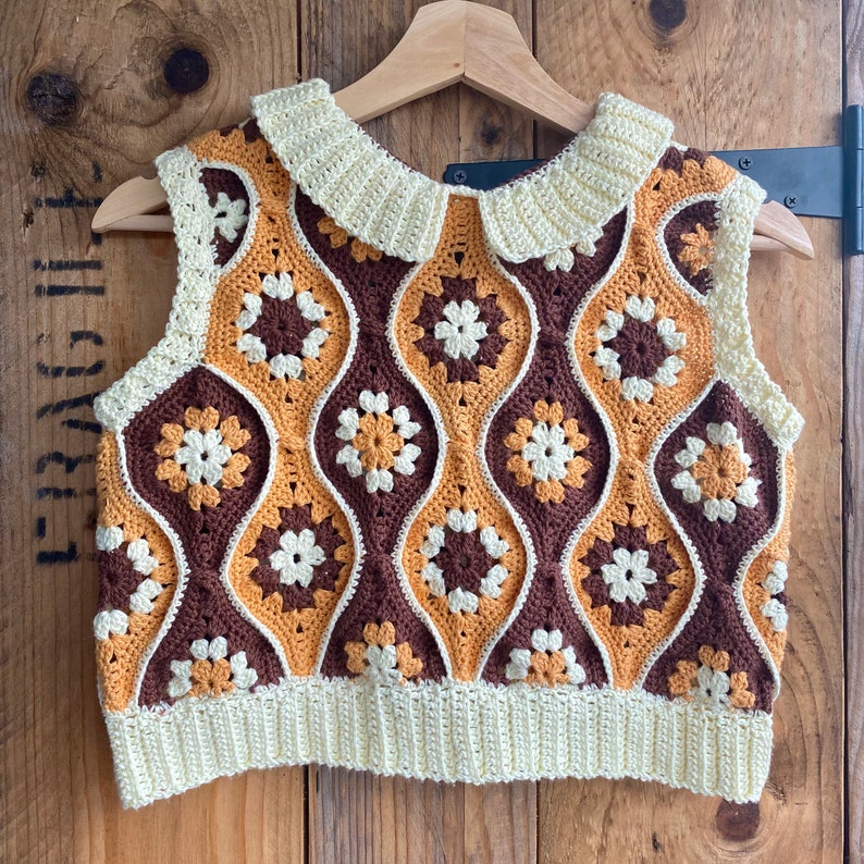 Pu&ograve; includere: Un top senza maniche all'uncinetto marrone, giallo e bianco con un colletto all'uncinetto bianco. Il top ha un motivo geometrico.