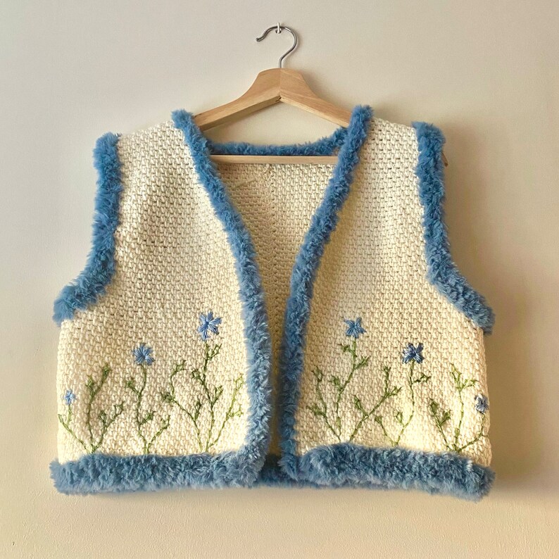 Pu&ograve; includere: Un gilet di uncinetto bianco con bordino in pelliccia sintetica blu e fiori blu ricamati.