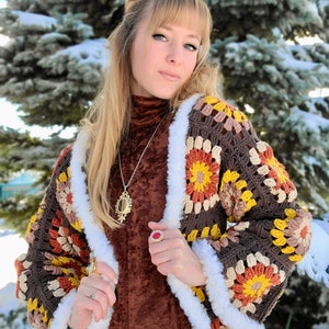 Pu&ograve; includere: Una donna indossa un cardigan di uncinetto marrone e bianco con un bordo di pelliccia. Indossa una camicia marrone a collo alto e una collana d'oro con un ciondolo a specchio. Indossa anche un anello rosso al dito.