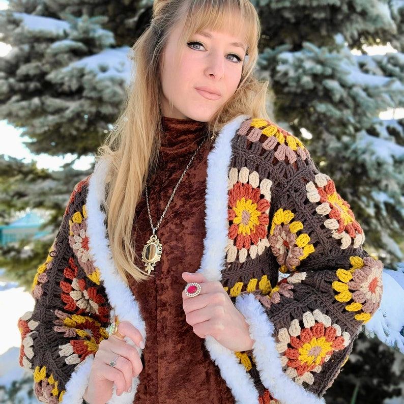 Pu&ograve; includere: Una donna indossa un cardigan di uncinetto marrone e bianco con un bordo di pelliccia. Indossa una camicia marrone a collo alto e una collana d'oro con un ciondolo a specchio. Indossa anche un anello rosso al dito.