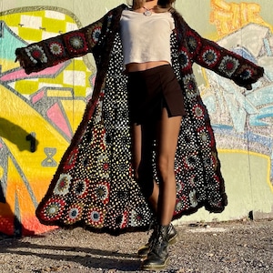Pu&ograve; includere: Un lungo cardigan in crochet nero con un motivo floreale colorato. Il cardigan &egrave; indossato sopra una canotta bianca e pantaloncini neri.