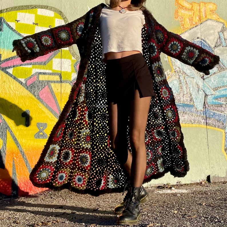 Pu&ograve; includere: Un lungo cardigan in crochet nero con un motivo floreale colorato. Il cardigan &egrave; indossato sopra una canotta bianca e pantaloncini neri.