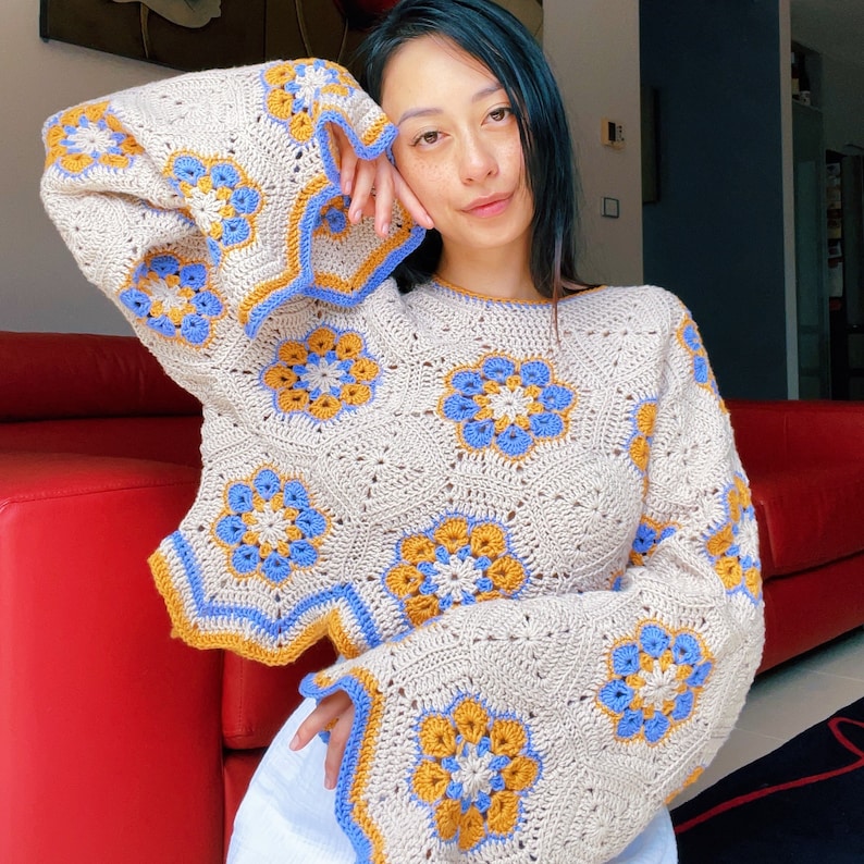 Pu&ograve; includere: Un maglione color crema lavorato a uncinetto con accenti floreali blu e gialli. Il maglione ha una vestibilit&agrave; ampia e maniche lunghe.