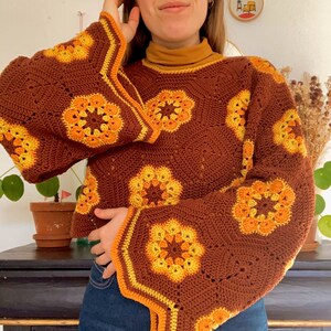 Pu&ograve; includere: Un maglione di uncinetto marrone con un design a maniche a campana. Il maglione presenta un motivo geometrico con accenti gialli e arancioni.