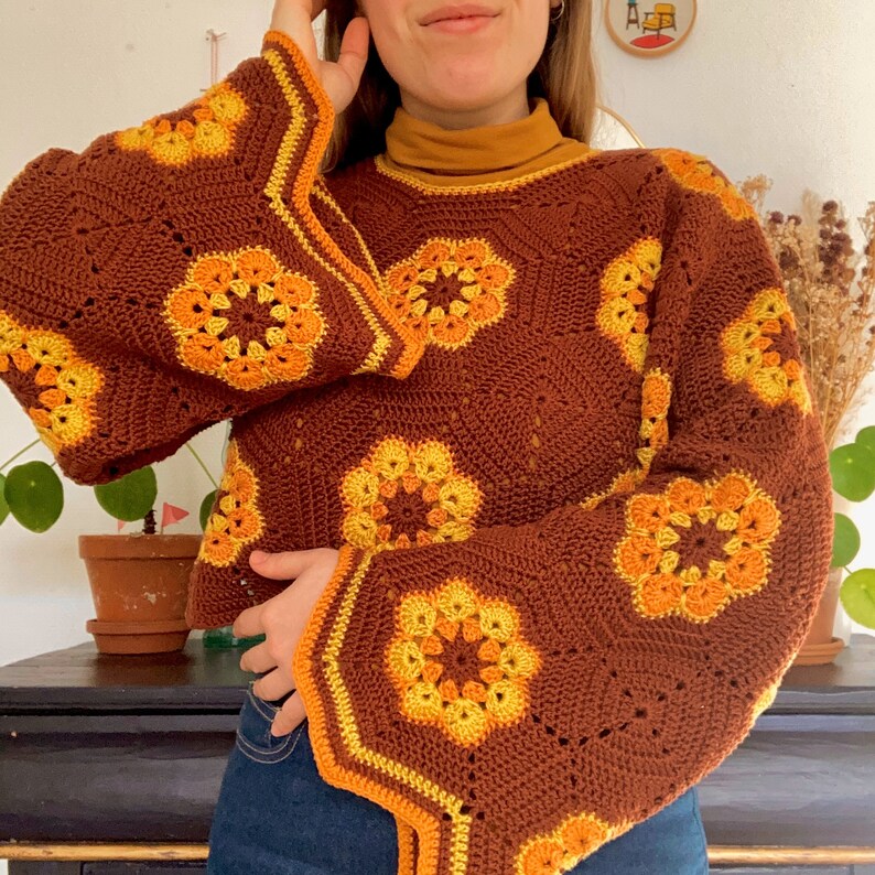 Pu&ograve; includere: Un maglione di uncinetto marrone con un design a maniche a campana. Il maglione presenta un motivo geometrico con accenti gialli e arancioni.