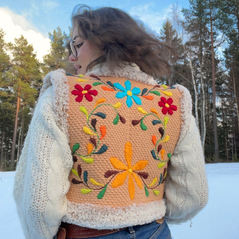 Pu&ograve; includere: Un gilet di maglia color crema con bordino in pelliccia sintetica marrone. Il gilet presenta fiori e viti ricamate colorate sul retro.