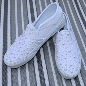 May include: A pair of white slip-on sneakers adorned with numerous faux pearls. The shoes have a classic design with a rounded toe and a rubber sole. The pearls are of varying sizes, adding texture and visual interest to the footwear.