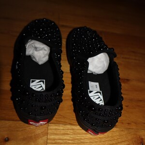Puede incluir: Un par de zapatillas negras sin cordones cubiertas de peque&ntilde;as cuentas oscuras. Los zapatos tienen una suela negra con un acento rojo cerca del tal&oacute;n y el logotipo de Vans en la plantilla.