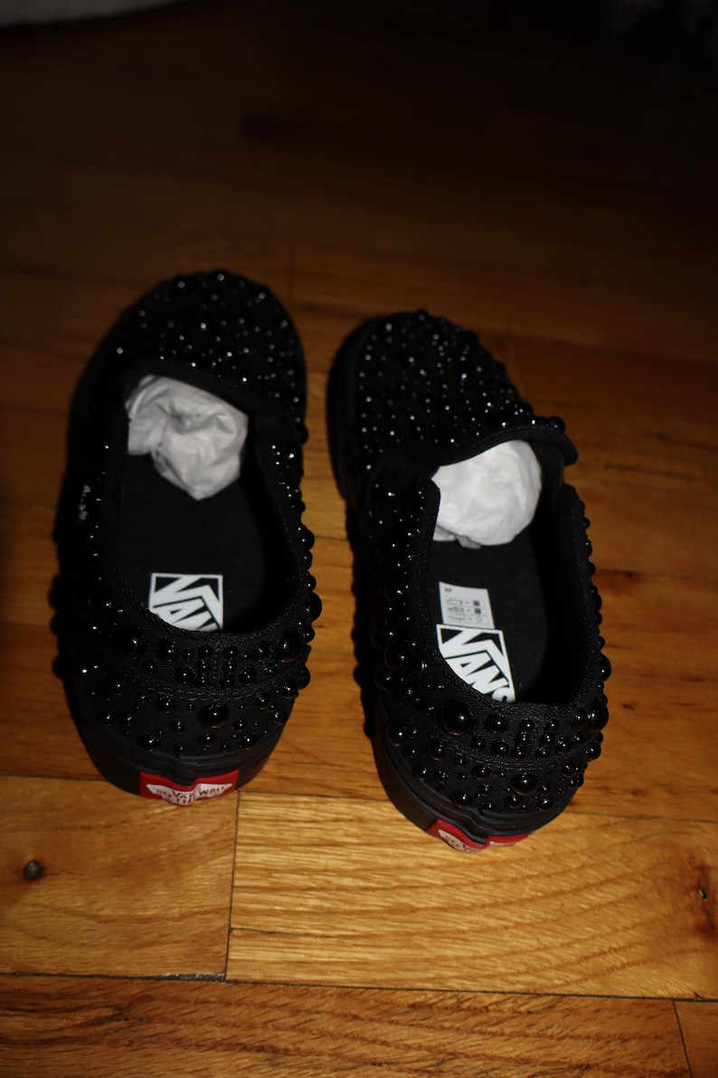 Puede incluir: Un par de zapatillas negras sin cordones cubiertas de peque&ntilde;as cuentas oscuras. Los zapatos tienen una suela negra con un acento rojo cerca del tal&oacute;n y el logotipo de Vans en la plantilla.