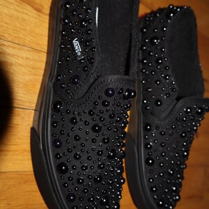 Puede incluir: Zapatillas negras sin cordones cubiertas de peque&ntilde;as decoraciones redondas y oscuras. Los zapatos tienen una suela negra y el logotipo de Vans es visible. Los zapatos se exhiben sobre una superficie de madera.