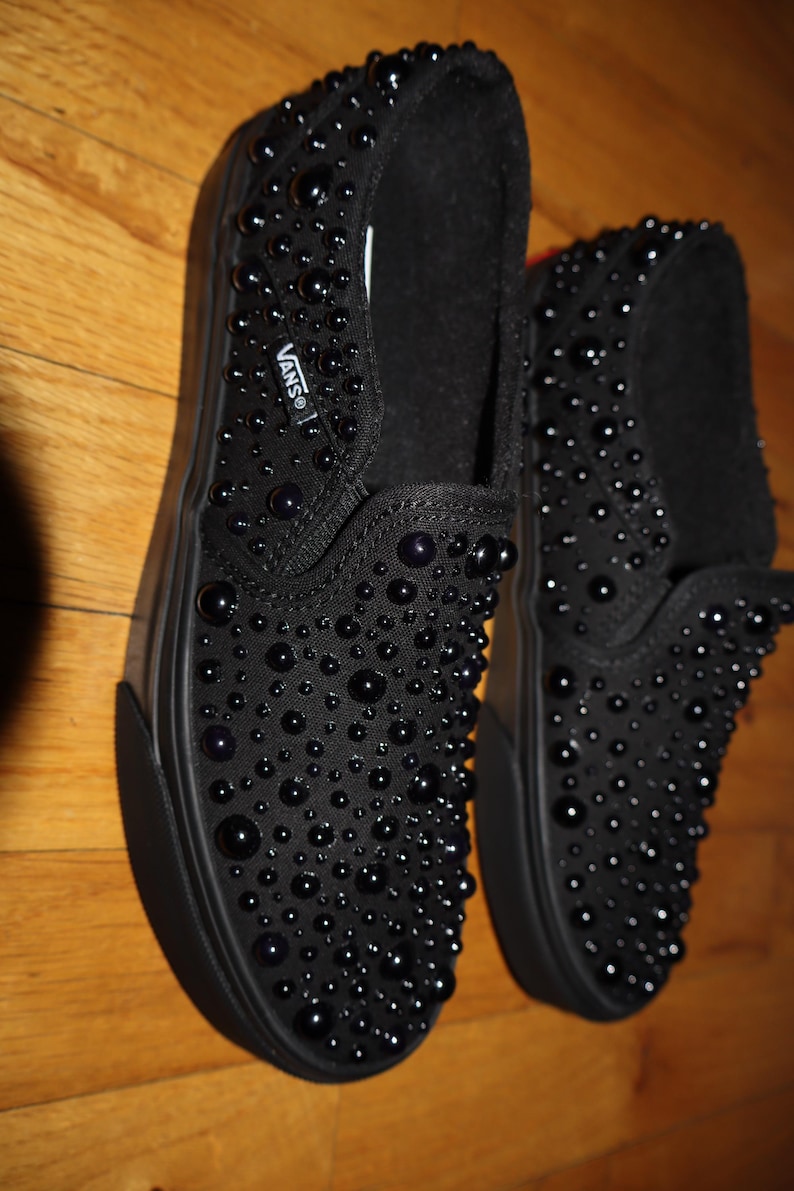 Puede incluir: Zapatillas negras sin cordones cubiertas de peque&ntilde;as decoraciones redondas y oscuras. Los zapatos tienen una suela negra y el logotipo de Vans es visible. Los zapatos se exhiben sobre una superficie de madera.