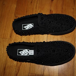 Puede incluir: Par de zapatillas negras sin cordones con dise&ntilde;o tachonado. Las zapatillas tienen el logotipo de Vans en blanco en la plantilla. Las zapatillas se muestran sobre un suelo de madera.