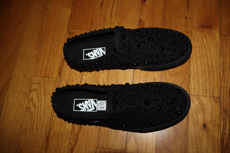 Puede incluir: Par de zapatillas negras sin cordones con dise&ntilde;o tachonado. Las zapatillas tienen el logotipo de Vans en blanco en la plantilla. Las zapatillas se muestran sobre un suelo de madera.