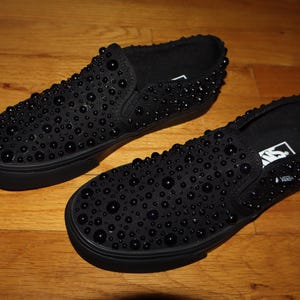 Puede incluir: Un par de zapatillas negras sin cordones cubiertas de peque&ntilde;os adornos redondos y oscuros. Los zapatos est&aacute;n sobre una superficie de madera. Las suelas y los ribetes de los zapatos tambi&eacute;n son negros. El logotipo de Vans es visible.