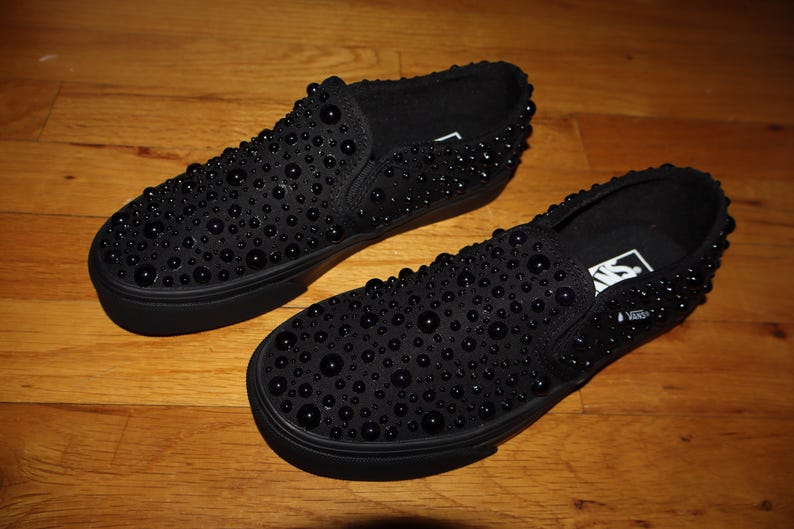 Puede incluir: Un par de zapatillas negras sin cordones cubiertas de peque&ntilde;os adornos redondos y oscuros. Los zapatos est&aacute;n sobre una superficie de madera. Las suelas y los ribetes de los zapatos tambi&eacute;n son negros. El logotipo de Vans es visible.