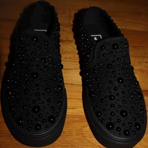 Puede incluir: Un par de zapatillas negras sin cordones adornadas con numerosas cuentas negras de varios tama&ntilde;os. Los zapatos tienen una suela negra s&oacute;lida y una puntera redondeada. La superficie de los zapatos est&aacute; cubierta de cuentas, creando una apariencia texturizada.