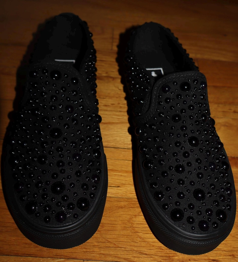 Puede incluir: Un par de zapatillas negras sin cordones adornadas con numerosas cuentas negras de varios tama&ntilde;os. Los zapatos tienen una suela negra s&oacute;lida y una puntera redondeada. La superficie de los zapatos est&aacute; cubierta de cuentas, creando una apariencia texturizada.