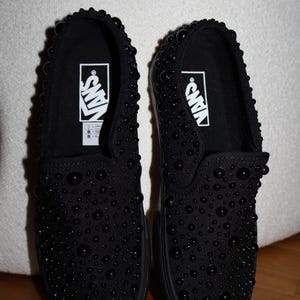 Puede incluir: Par de zapatillas negras sin cordones con un dise&ntilde;o tachonado. Los zapatos presentan el logotipo de Vans en blanco en el interior. Las zapatillas se exhiben sobre una superficie de madera.